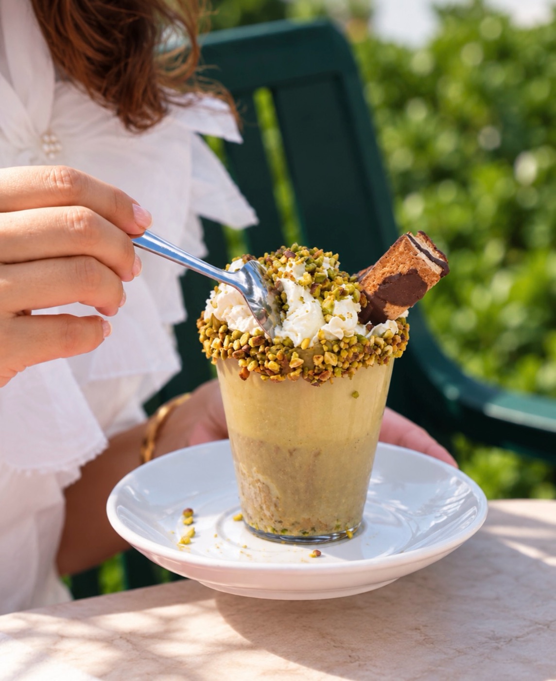 A pistachio dessert in a glass, topped with crumbled pistachio and a wafer, on a terrace table