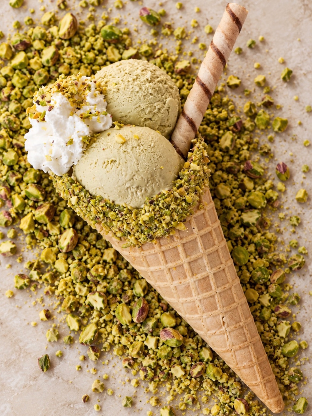 A waffle cone with two scoops of pistachio gelato and whipped cream on crushed pistachios